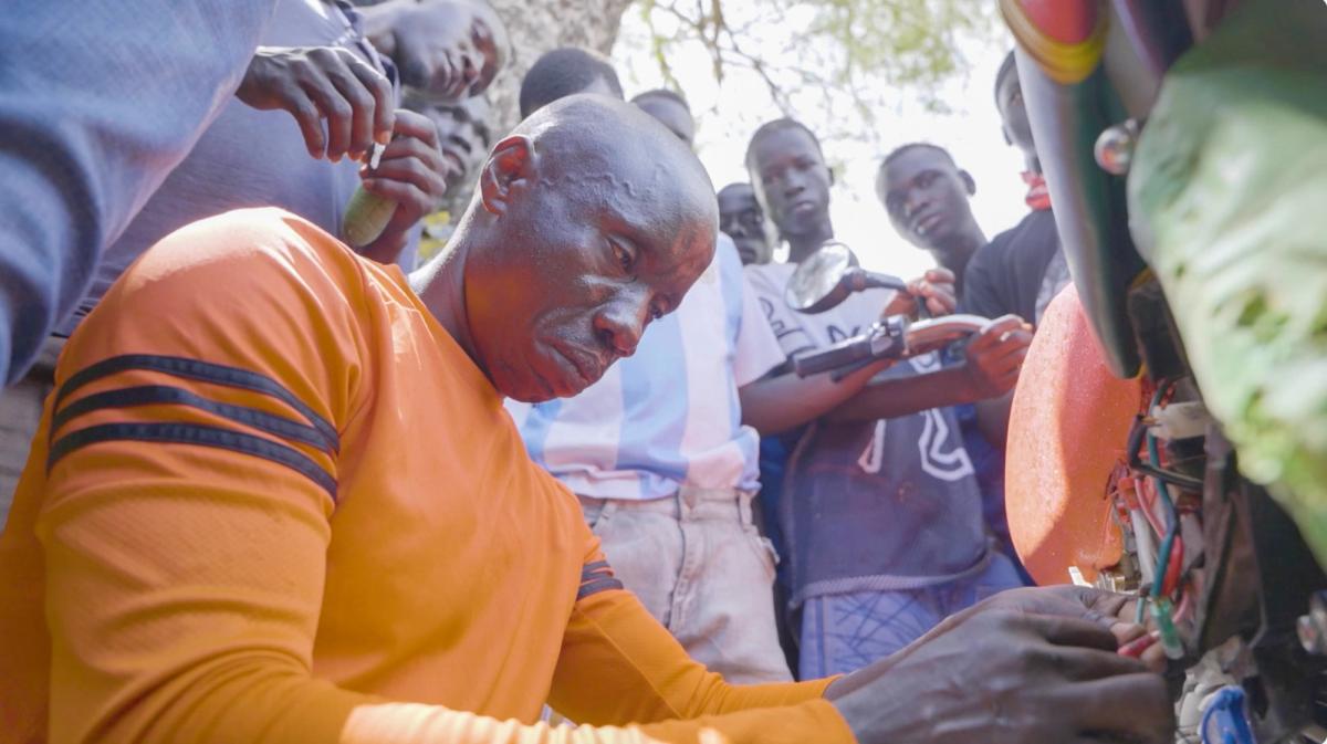 A man in an orange long-sleeve shirt concentrates while repairing electrical wiring on a motorcycle. Several young men stand closely behind him, watching intently, with one holding a mango and another leaning on the motorcycle’s handlebars. The group is gathered outdoors under a tree, with sunlight filtering through the leaves.